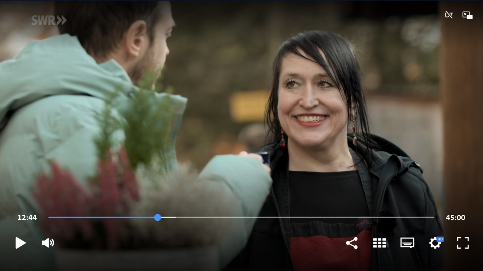 Lächelnde Frau spricht mit einem Mann, im Vordergrund stehen Pflanzen.