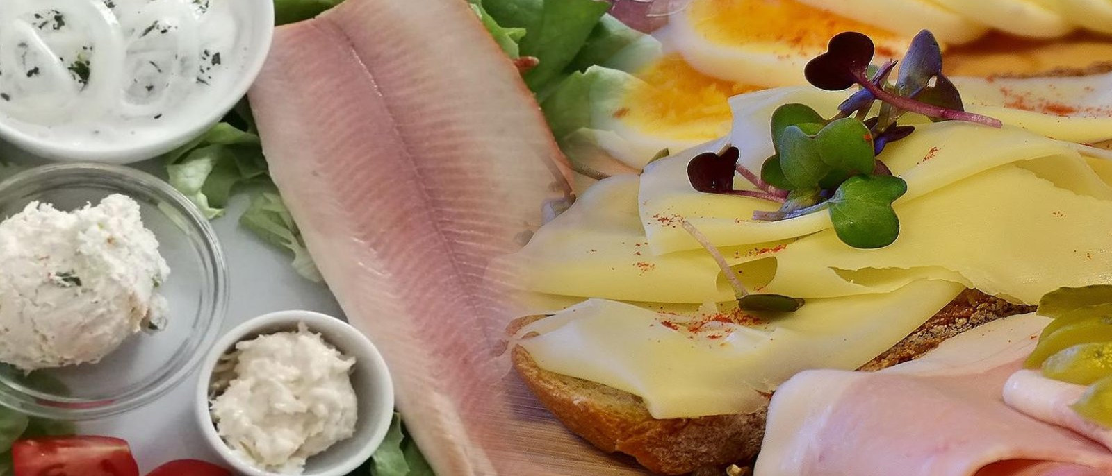 Geräucherter Fisch mit Saucen und Gemüse neben Brot mit Aufschnitt, Käse und Eiern.