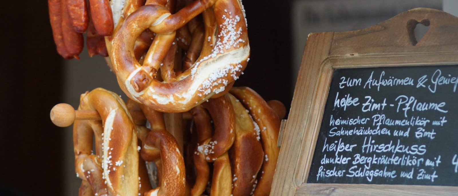 Brezeln und Würstchen hängen an einem Stand, mit einer Tafel mit Beschriftungen daneben.