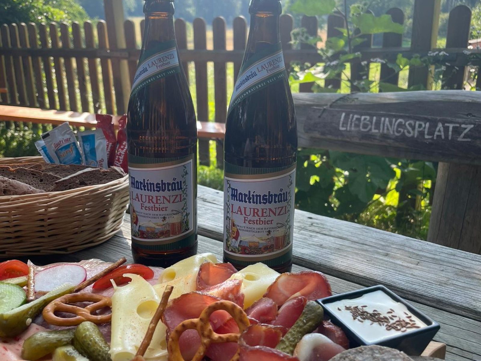 Brotzeitplatte mit Aufschnitt und Käse auf einem Tisch, zwei Bierflaschen daneben, im Freien.