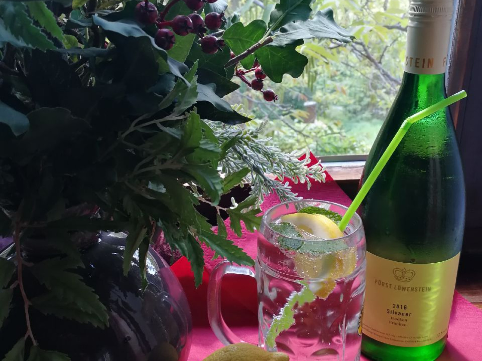 Ein Glas mit Zitronenwasser, eine Weinflasche und Zitronen neben einem Blumenstrauß vor einem Fenster.