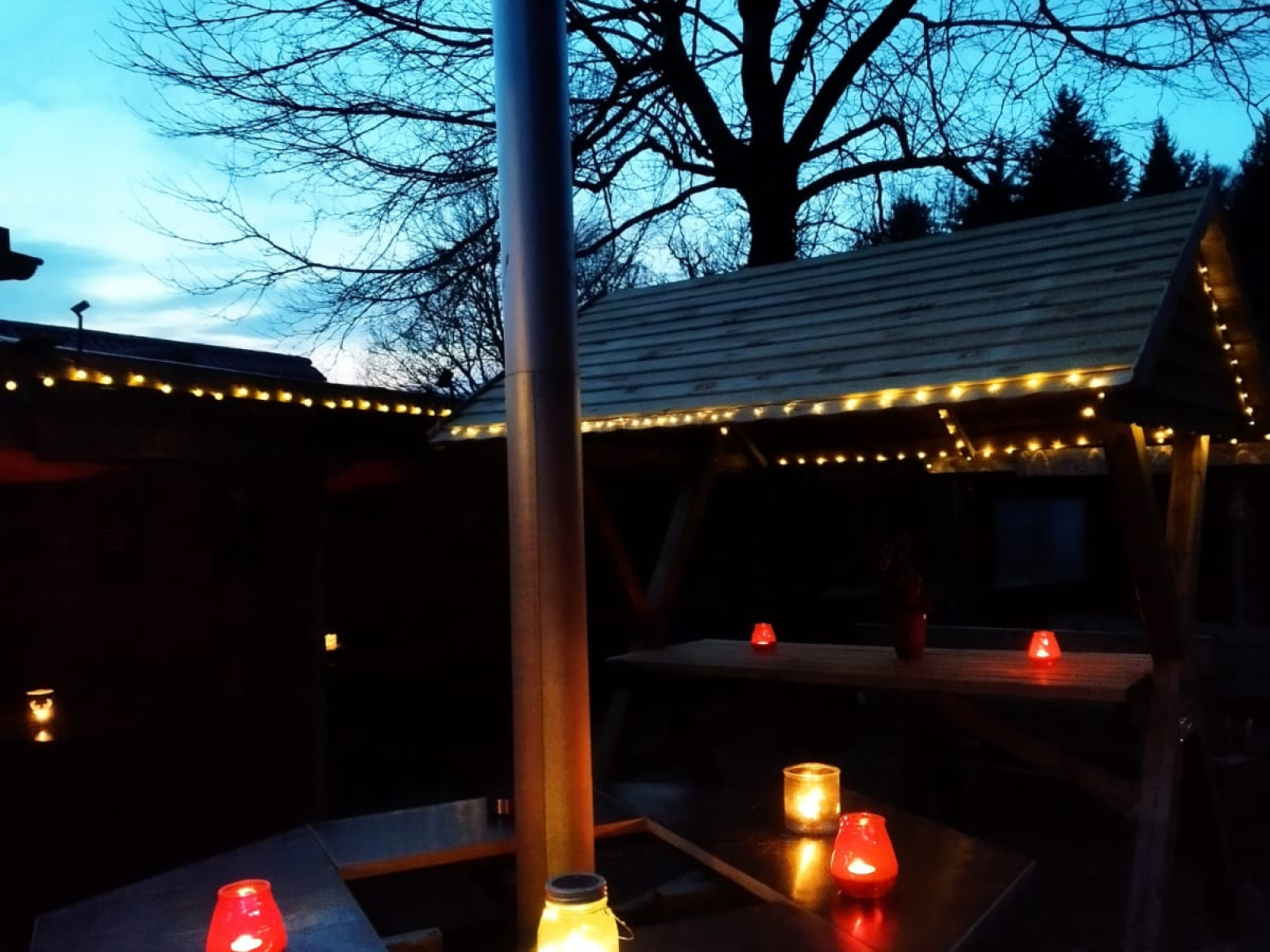 Außenbereich mit Tisch und Kerzenlichtern, beleuchtet von Lichterketten, bei Dämmerung.
