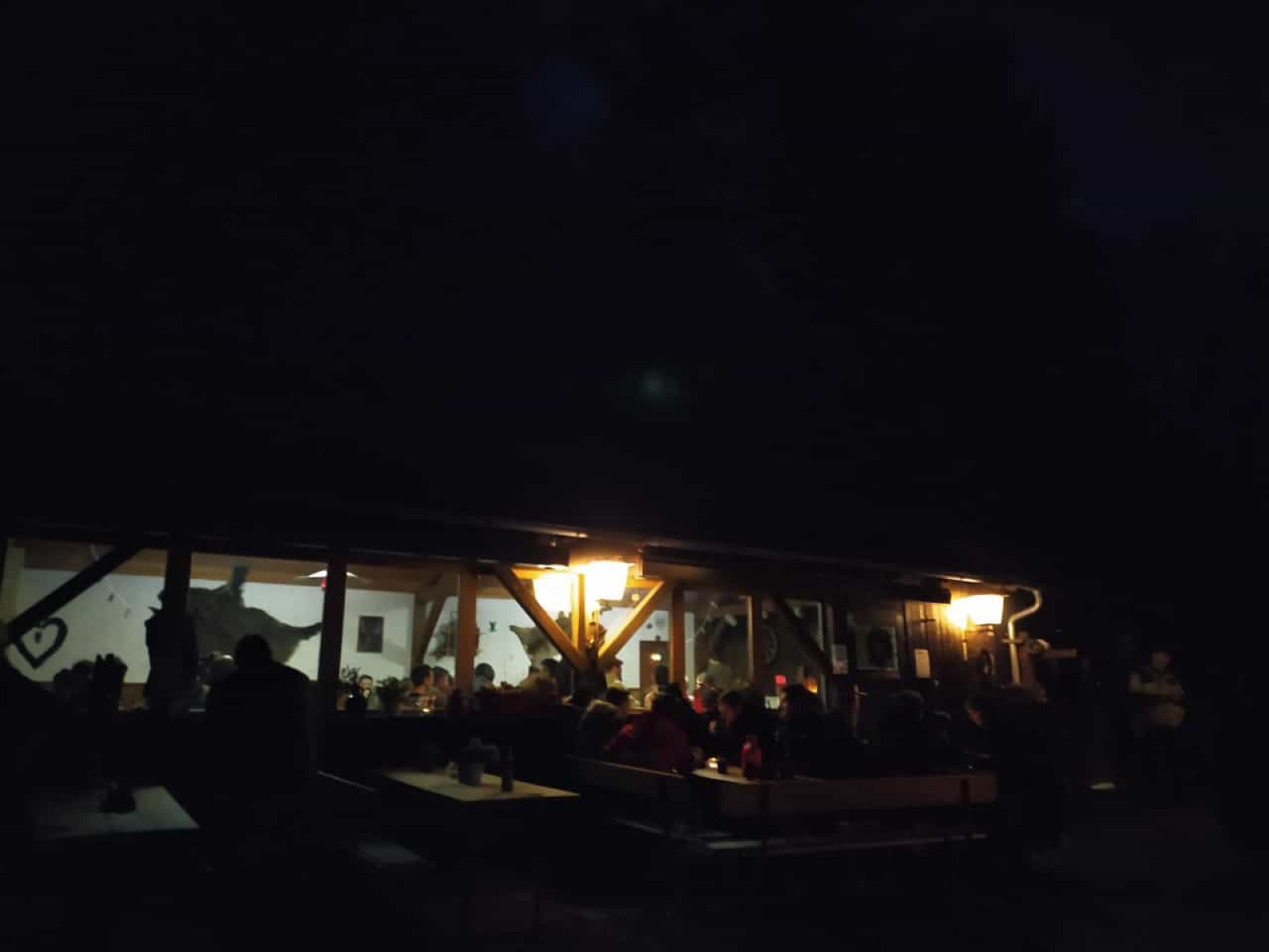 Abendszene mit Menschen in einem beleuchteten Gebäude, umrissene Gestalten draußen im Dunkeln.