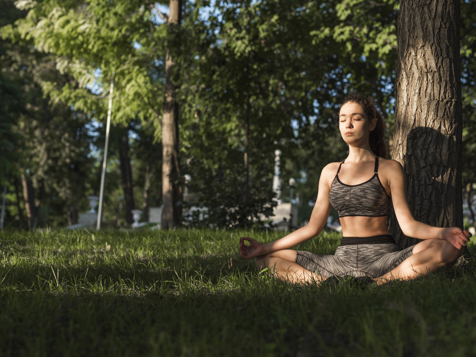 junge Frau in Yoga Pose vor einem Baumstamm, im gras sitzend