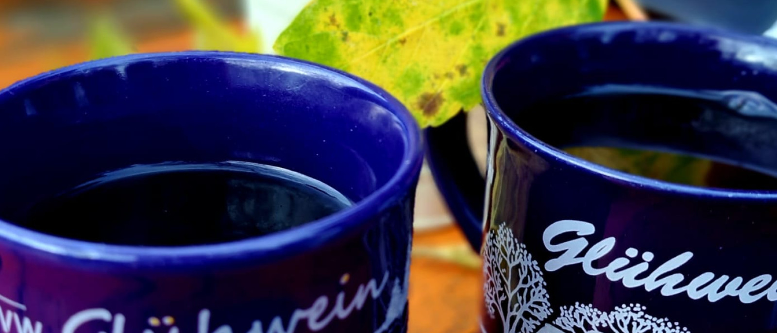 Zwei blaue Tassen mit Glühwein und ein großes gelbes Herbstblatt auf Holztisch.